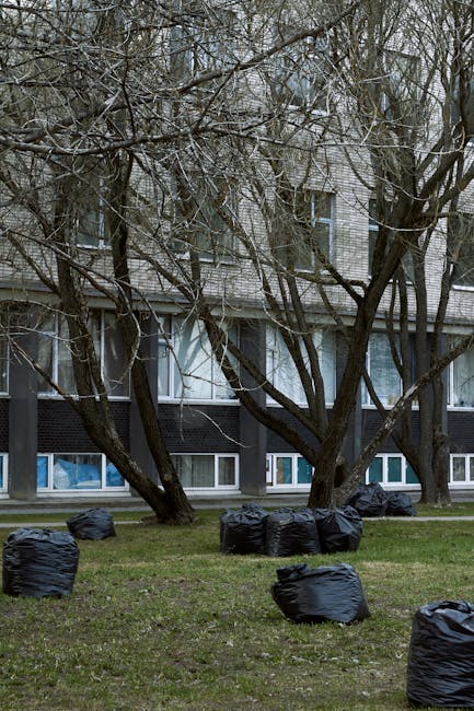 Two large black trash bags filled with waste are positioned on the pavement beside a black metal fence, which has vertical bars and a diamond-patterned lattice section. One bag appears to be partially open, revealing objects inside, while the other is tightly knotted. The bags are situated near the edge of the sidewalk, with a curb visible in the foreground. Behind the fence, leafy bushes or trees provide a dense green backdrop, contrasting with the dark shadows cast on the scene. The environment suggests an urban or suburban setting where private waste collection or on-site rubbish clearance may be taking place, and the presence of Waste Collection Kentish Town could imply a service for non-curbside rubbish removal. The lighting is subdued, possibly due to overcast weather or early evening conditions, emphasizing the textures of the plastic bags and the surrounding environment. This image exemplifies typical street-side rubbish awaiting collection or disposal, aligned with private waste management services rather than local authority collection.