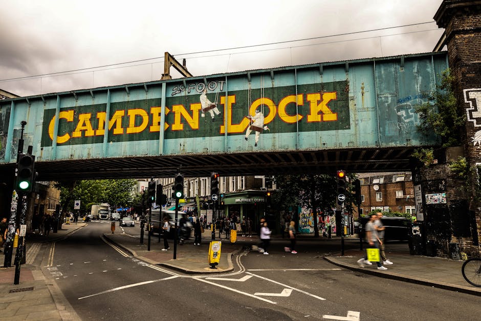 A large, weathered metal bridge structure painted in faded turquoise and rust tones, bearing the bold yellow text 'CAMDEN LOCK' with a silhouette of a person hanging from a rope or cable, situated above a busy urban intersection. The underside of the bridge reveals multiple rust streaks and peeling paint, indicating age and exposure to the elements. The scene below includes a traffic light with a green signal, several pedestrians crossing the road, some walking and others waiting. To the left, a black lamppost and signage are visible, with additional street fixtures and bollards guiding vehicle and pedestrian flow. In the background, a mixture of residential and commercial buildings, some partially covered in graffiti or posters, frame the scene. The environment is overcast with grey clouds, contributing to a muted and somewhat industrial atmosphere. This urban setting, with its aged infrastructure and active street life, visually connects to alternative waste handling or on-site clearance services that might operate in busy city areas, as exemplified by Waste Collection Kentish Town’s scope of rubbish removal in such contexts.