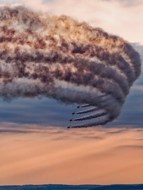 Four jet aircrafts fly in close formation, leaving behind long, dense trails of grey smoke that spread across the sky during sunset. The aircraft are small and dark, visible against the vibrant, warm-toned sky with shades of orange, pink, and purple. The smoke trails curve slightly, indicating rapid movement and precision flying. Below, a distant horizon line is visible, with a flat landscape that appears dark and unobstructed. The scene captures the dynamic motion of the jets and the dramatic contrast of their smoke trails against the colorful sky, which could relate to aerial displays or high-performance flight activities. This image, although focused on an aviation event, exemplifies clearing large debris or managing complex waste removal tasks with specialized equipment, akin to the precision required in industrial or large-scale rubbish disposal handled by companies like Waste Collection Kentish Town.