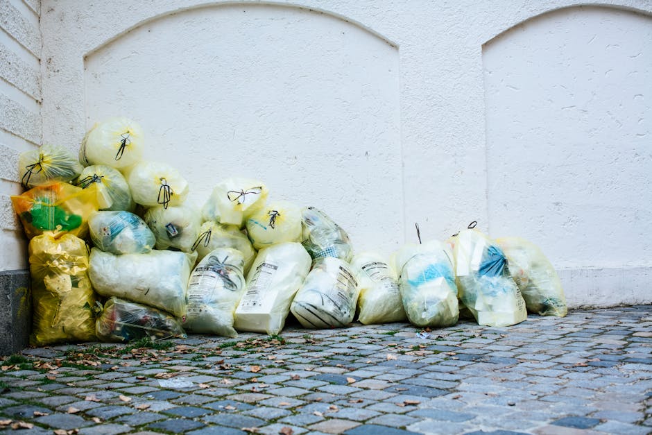 A collection of yellow and white plastic rubbish bags filled with waste, stacked against a white textured wall on a cobblestone pavement. The bags appear to contain general household waste, with some partly transparent to reveal contents such as paper and plastic. Several bags are tied with black drawstrings, while others are closed with twist ties or knotted ties. The scene is outdoors, under natural lighting, with a corner of a grey brick wall visible on the left. The ground is uneven with small pebble-like stones and some scattered leaves and debris around the bags. This setup suggests an informal waste collection point, possibly managed by a private waste removal service like Waste Collection Kentish Town, and reflects a typical scenario where bulkier rubbish or overflow waste might be temporarily stored prior to removal or disposal, supporting the context of alternative or private rubbish collection methods in urban environments.