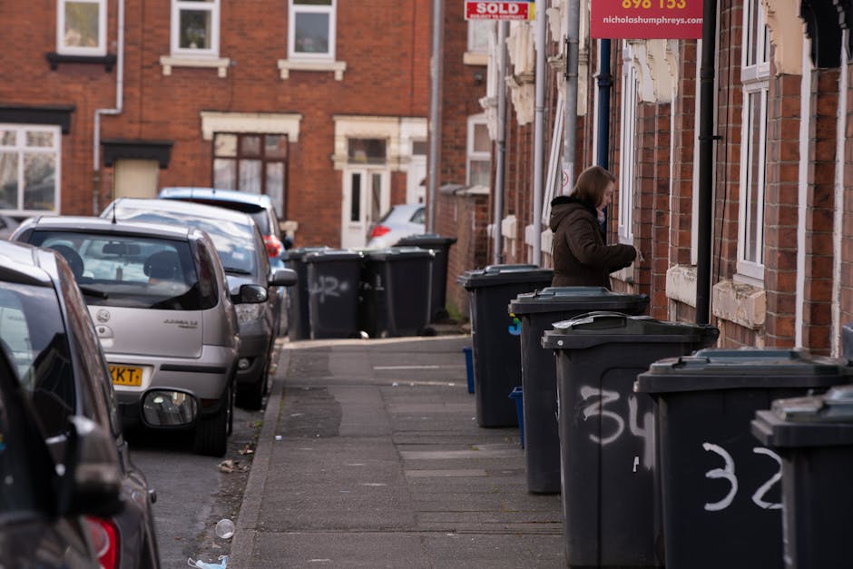 A row of dark grey wheelie bins with closed lids is lined up along the pavement outside a brick residential building. The bins are positioned close to the building’s wall, which features multiple white-framed windows and brick detailing. One woman is standing next to a bin, holding items in her hand, appearing to be about to dispose of waste or organize the bins. Several parked cars, including a silver hatchback and a darker vehicle, are situated along the street, occupying parking spaces adjacent to the sidewalk. The background reveals more brick buildings with similar architectural features, and additional bins placed along the same section of pavement, suggesting regular waste disposal activity. The scene is illuminated by natural daylight, and some graffiti is visible on the lower part of the bins. The overall environment depicts a typical residential street scene with waste management elements consistent with private rubbish collection services, as operated by Waste Collection Kentish Town.