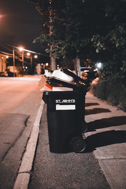 A row of dark grey wheelie bins with closed lids is lined up along the pavement outside a brick residential building. The bins are positioned close to the building’s wall, which features multiple white-framed windows and brick detailing. One woman is standing next to a bin, holding items in her hand, appearing to be about to dispose of waste or organize the bins. Several parked cars, including a silver hatchback and a darker vehicle, are situated along the street, occupying parking spaces adjacent to the sidewalk. The background reveals more brick buildings with similar architectural features, and additional bins placed along the same section of pavement, suggesting regular waste disposal activity. The scene is illuminated by natural daylight, and some graffiti is visible on the lower part of the bins. The overall environment depicts a typical residential street scene with waste management elements consistent with private rubbish collection services, as operated by Waste Collection Kentish Town.