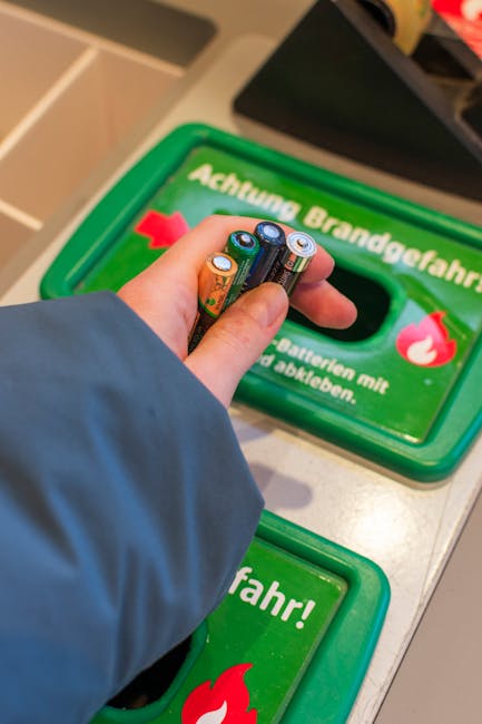 A person wearing a blue jacket is inserting used batteries into a green recycling bin designated for hazardous waste disposal. The person's hand is holding three AA batteries, with the positive terminals visible; the batteries have metallic tops and colored labels. The recycling bin has a bright green lid with a prominent red flame icon and text in German, indicating it is for disposing of batteries with a specific warning message. The background shows a countertop or table surface, possibly in a public or retail environment, with additional waste collection containers nearby. The scene emphasizes the proper handling of small electrical waste and the use of designated collection points, aligning with independent waste disposal practices offered by companies like Waste Collection Kentish Town. Lighting is bright, highlighting the textures of the batteries and the smooth plastic finish of the recycling container, conveying a clear view of battery recycling procedures without assumptions beyond the visible details.