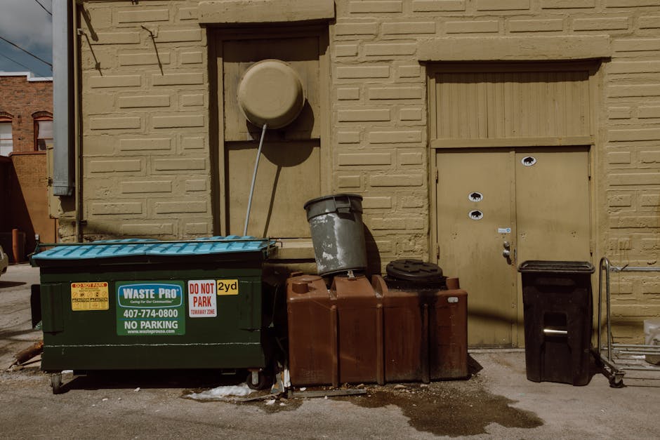 An outdoor scene in an urban alleyway features a variety of waste disposal items positioned against a yellow brick wall. In the foreground, a large dark green waste container with a blue lid displays a waste collection company label, including contact details and no parking signage, indicating it is used for commercial or private rubbish disposal. To its right, there is a tall, rust-colored, rectangular plastic bin, partially filled with waste, with weathered surfaces visible. Adjacent to it, a smaller black bin with a closed lid stands upright, accompanied by a metal trolley with a handle, typically used for transporting rubbish. Behind these containers, a grey trash bin with a lid is tilted slightly, resting against a wooden panel attached to the brick wall. Above these items, a large circular grey cover, possibly for a vent or utility access point, is mounted on the wall, with a metal pipe extending diagonally downward from its center. The background reveals portions of other brick buildings and a clear sky, with shadows cast by the objects suggesting bright daylight. The scene reflects a typical private waste collection or rubbish disposal setup in an urban environment, managed by Waste Collection Kentish Town, intended for the efficient removal of household or commercial refuse from the designated premises.