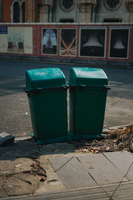A person wearing a blue jacket is inserting used batteries into a green recycling bin designated for hazardous waste disposal. The person's hand is holding three AA batteries, with the positive terminals visible; the batteries have metallic tops and colored labels. The recycling bin has a bright green lid with a prominent red flame icon and text in German, indicating it is for disposing of batteries with a specific warning message. The background shows a countertop or table surface, possibly in a public or retail environment, with additional waste collection containers nearby. The scene emphasizes the proper handling of small electrical waste and the use of designated collection points, aligning with independent waste disposal practices offered by companies like Waste Collection Kentish Town. Lighting is bright, highlighting the textures of the batteries and the smooth plastic finish of the recycling container, conveying a clear view of battery recycling procedures without assumptions beyond the visible details.
