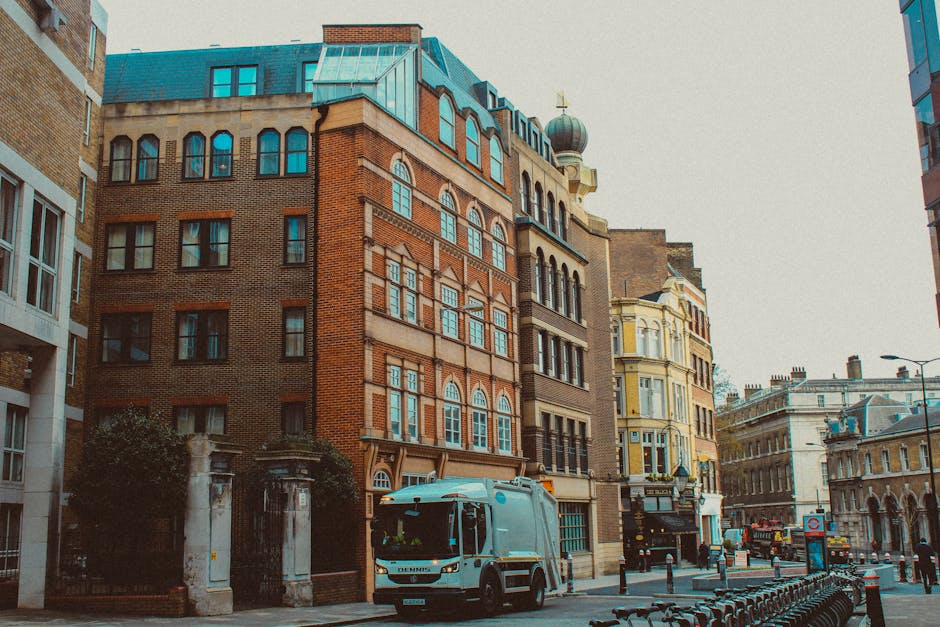 A street scene featuring a city block with historic brick buildings and ornate architectural details, including large arched windows and decorative facades. In the foreground, a waste collection truck with a blue cab is parked on the pavement, partially obscuring an entrance gate with stone pillars. The truck appears to be engaged in rubbish collection or transportation, aligning with private waste disposal services. To the right, a row of bicycle racks is visible alongside the curb, and further down the street, additional vehicles and street furniture, such as streetlights and bollards, can be seen. The buildings exhibit a mixture of red, beige, and cream-colored facades, with some featuring domed towers and intricate cornices, under a clear sky with natural daylight illuminating the scene. The environment suggests an urban setting where independent waste collection services may operate alongside public infrastructure, emphasizing the importance of efficient rubbish removal for city management. Waste Collection Kentish Town’s service relevance is subtly reflected through the depiction of commercial vehicle presence within a densely built historic area, underlining the professional handling of waste in such environments.