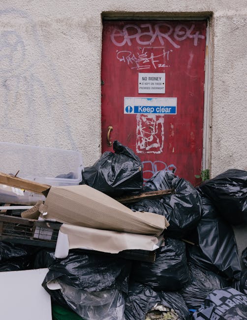 A large pile of black trash bags, some containing miscellaneous waste, is stacked against a textured, off-white exterior wall beneath a red metal door with visible graffiti and stickers. The bags are packed tightly, with some torn or slightly flattened, revealing bits of cardboard and other refuse. In front of the pile, there are loose pieces of discarded items, including a flattened cardboard box and a piece of foam or upholstery material. The red metal door, secured within a rough concrete frame, features several stickers, including one clearly reading 'Keep clear' and another with the message 'No money' printed on a white background, along with graffiti tags in white and pink spray paint. The scene suggests an informal or alternative waste collection area, illustrating the challenges of managing private rubbish disposal in an urban environment, possibly indicating a need for professional rubbish removal services like those offered by Waste Collection Kentish Town, especially for on-site clearance or handling non-household waste outside regular municipal collection points.
