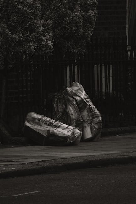 Two large black plastic bags filled with waste material are tied at the top and placed on a paved sidewalk in front of a tall wooden fence with vertical slats. The bags appear to contain household or garden rubbish and are positioned close to the curb, partially overlapping each other. Behind the fence, dense bushes or trees with dark, leafy foliage are visible, creating a shaded background. The scene is captured outdoors in natural light, with the pavement and road surface in the foreground. The environment suggests an informal rubbish collection or disposal area, possibly managed by private waste services such as Waste Collection Kentish Town, offering alternative waste handling outside local authority collection methods.