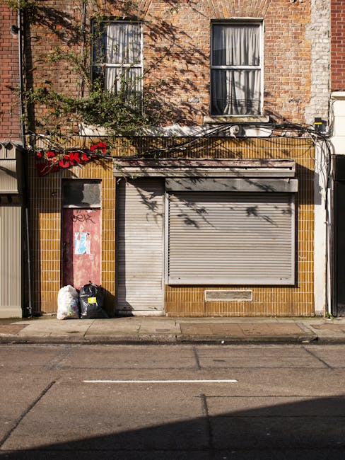 Two large black plastic bags filled with waste material are tied at the top and placed on a paved sidewalk in front of a tall wooden fence with vertical slats. The bags appear to contain household or garden rubbish and are positioned close to the curb, partially overlapping each other. Behind the fence, dense bushes or trees with dark, leafy foliage are visible, creating a shaded background. The scene is captured outdoors in natural light, with the pavement and road surface in the foreground. The environment suggests an informal rubbish collection or disposal area, possibly managed by private waste services such as Waste Collection Kentish Town, offering alternative waste handling outside local authority collection methods.