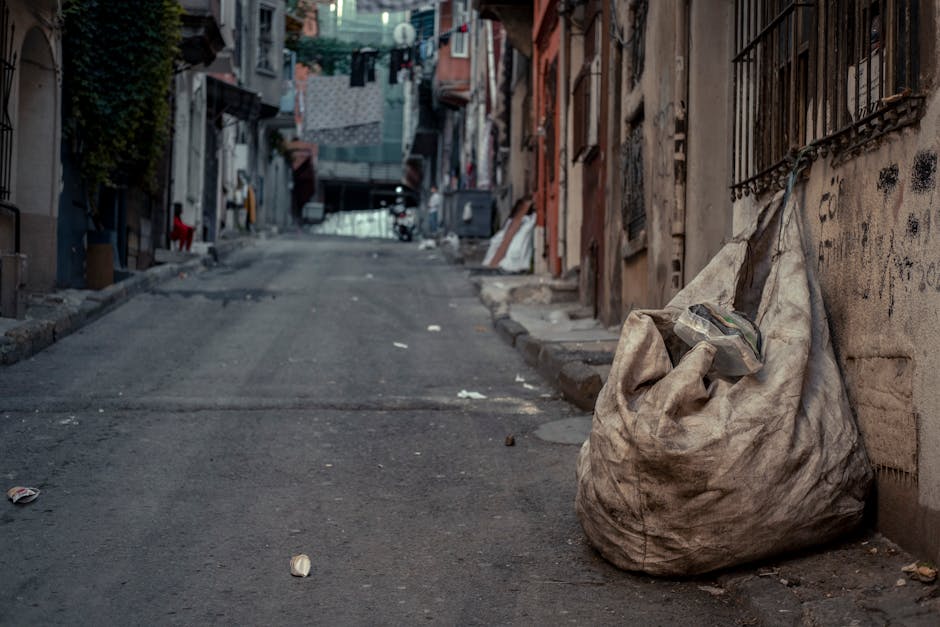 A large, overfilled beige rubbish sack made of durable woven material is positioned on the right side of a narrow, uneven city street, leaning against a weathered brick and plaster wall. The sack appears worn, dirty, and bulging with waste, with a partially visible black garbage bag or plastic wrapping protruding from the top. The street surface is rough and cracked, with scattered pieces of litter and small debris strewn across its dark, uneven asphalt. In the background, residential buildings with faded facades and barred windows line both sides of the street, some with laundry hanging on clotheslines and others with makeshift repairs. A metal railing, possibly covering a window or door, is visible on the right side, and faint graffiti markings can be seen on parts of the wall. The scene is lit by natural, diffuse daylight, creating a muted atmosphere that highlights the urban environment and the need for waste collection or disposal, subtly connecting to the services offered by Waste Collection Kentish Town for managing rubbish in such areas.