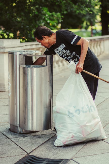 A middle-aged woman with short dark hair, wearing a black t-shirt with white text on the front, is seen discarding rubbish into a modern stainless steel litter bin situated on a paved sidewalk. She is holding a large white garbage bag in her left hand and using her right hand to empty its contents into the top of the bin, which features a vertical slit opening designed for waste disposal. The scene is set outdoors on a bright day, with green leafy trees and neatly maintained shrubbery in the background, providing a natural environment typical of urban park areas or pedestrian-friendly streets. The pavement is composed of large rectangular stone slabs, and the stainless steel bin has a reflective surface, contrasting with the darker tones of her clothing. The overall setting suggests an example of independent rubbish collection or waste management, with Waste Collection Kentish Town focused on efficient on-site clearance of refuse from public spaces into designated waste receptacles, supporting local waste disposal needs in the community.