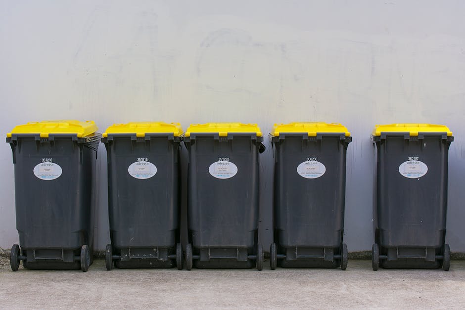 A large, overfilled beige rubbish sack made of durable woven material is positioned on the right side of a narrow, uneven city street, leaning against a weathered brick and plaster wall. The sack appears worn, dirty, and bulging with waste, with a partially visible black garbage bag or plastic wrapping protruding from the top. The street surface is rough and cracked, with scattered pieces of litter and small debris strewn across its dark, uneven asphalt. In the background, residential buildings with faded facades and barred windows line both sides of the street, some with laundry hanging on clotheslines and others with makeshift repairs. A metal railing, possibly covering a window or door, is visible on the right side, and faint graffiti markings can be seen on parts of the wall. The scene is lit by natural, diffuse daylight, creating a muted atmosphere that highlights the urban environment and the need for waste collection or disposal, subtly connecting to the services offered by Waste Collection Kentish Town for managing rubbish in such areas.
