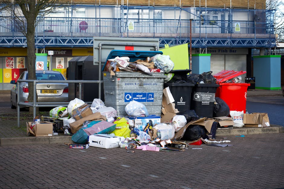 A large collection of mixed household waste and recyclable materials is piled up on a paved urban street at a designated waste collection point. The clutter includes cardboard boxes, paper bags, plastic shopping bags, and various wrapped items, some torn or spilled onto the ground. Several black, green, and red rubbish bins, some overflowing with waste, are positioned around the pile, with lids either open or partially ajar. Behind the waste collection site, a low metal railing separates the rubbish area from the adjacent parking space, where a grey car with a yellow license plate is parked. In the background, there is a storefront with brightly coloured signage and large windows, partially obscured by a tree with leafless branches, indicating potentially colder weather. A building with a blue scaffolding structure suggests ongoing maintenance or renovations. The lighting appears natural, consistent with daylight hours, highlighting the cluttered scene and the textured surfaces of the waste materials and urban surroundings. This scene reflects an instance of outside rubbish disposal, which may be addressed by services such as Waste Collection Kentish Town, specialising in rubbish collection and waste management solutions in the local area.