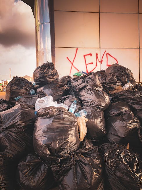 A large pile of black rubbish bags filled with waste materials is stacked against a modern tiled wall with a graffiti tag spelling 'XEND' in red spray paint. Some of the bags are torn or partially open, revealing plastic bottles and other debris inside. The scene appears to be outdoors, possibly on a pavement or in an alleyway, with urban buildings visible in the background under a cloudy sky. The waste bags are close to a reflective metallic surface, likely part of a building facade, and are arranged in a disorderly manner. The presence of plastic bottles among the waste indicates typical household or commercial refuse, and the overall scene highlights the need for waste collection services such as those offered by Waste Collection Kentish Town for effective on-site clearance and private rubbish disposal options.