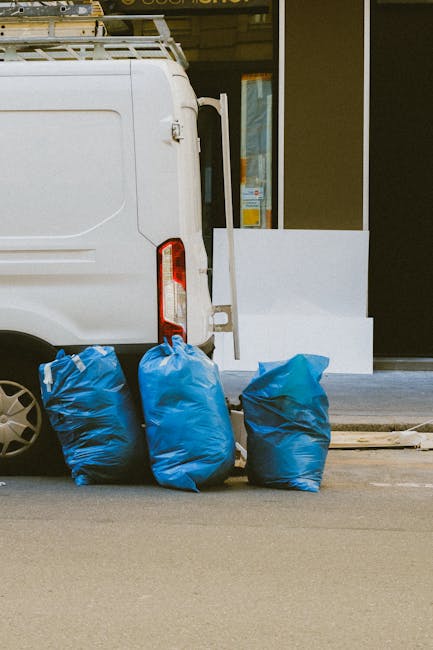Two large black plastic bags filled with waste material, tightly knotted at the top, are placed on the curbside pavement near a dark metal fence and dense foliage in the background. The bags appear to contain household or garden refuse, with visible crumpled and uneven surfaces, and some partially covered with printed branding or labels. The environment looks like an urban street scene, possibly in the late evening or at night, with minimal lighting casting shadows on the pavement and surroundings. This image illustrates an instance of private rubbish disposal, which waste collection services like Waste Collection Kentish Town might handle through alternative waste handling or on-site clearance in non-official waste collection contexts, reflecting the need for professional rubbish removal solutions.